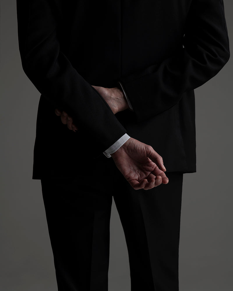 Person wearing a black suit with hands clasped in front on a gray background